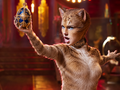 Fictional theatrical illustration of a feline performer holding an ornate blue-and-gold jeweled egg beneath warm stage lights.